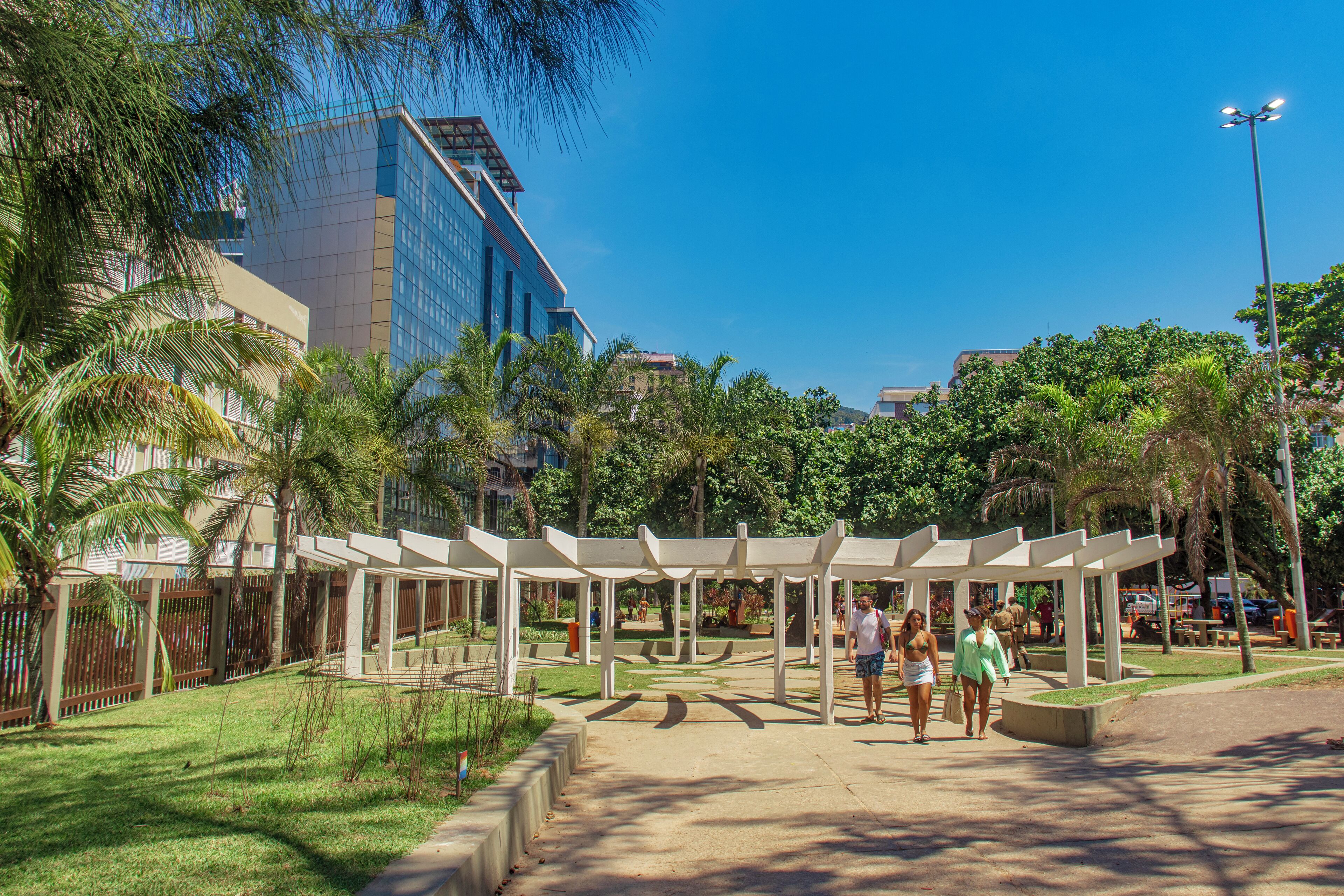 Um parque com um pérgola branca, palmeiras e um edifício moderno ao fundo.