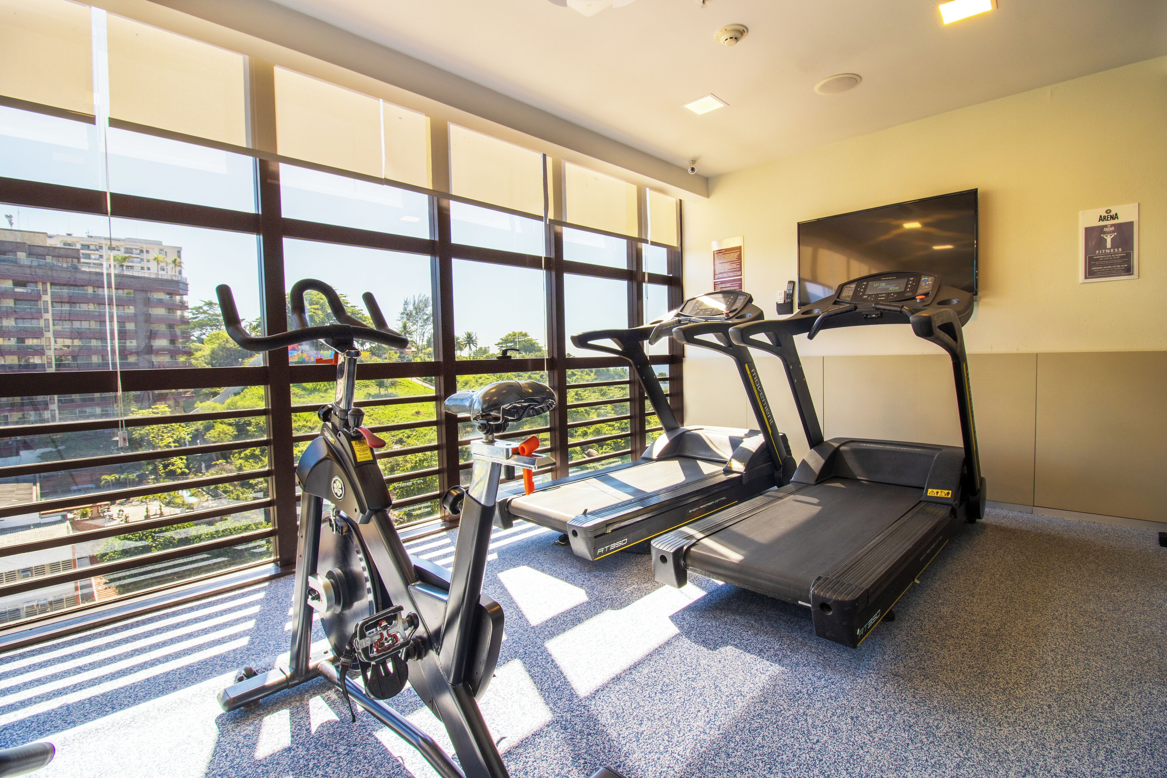 Ein Fitnessraum mit zwei Laufbändern und einem Hometrainer, große Fenster mit Blick auf Gebäude und Grünflächen.