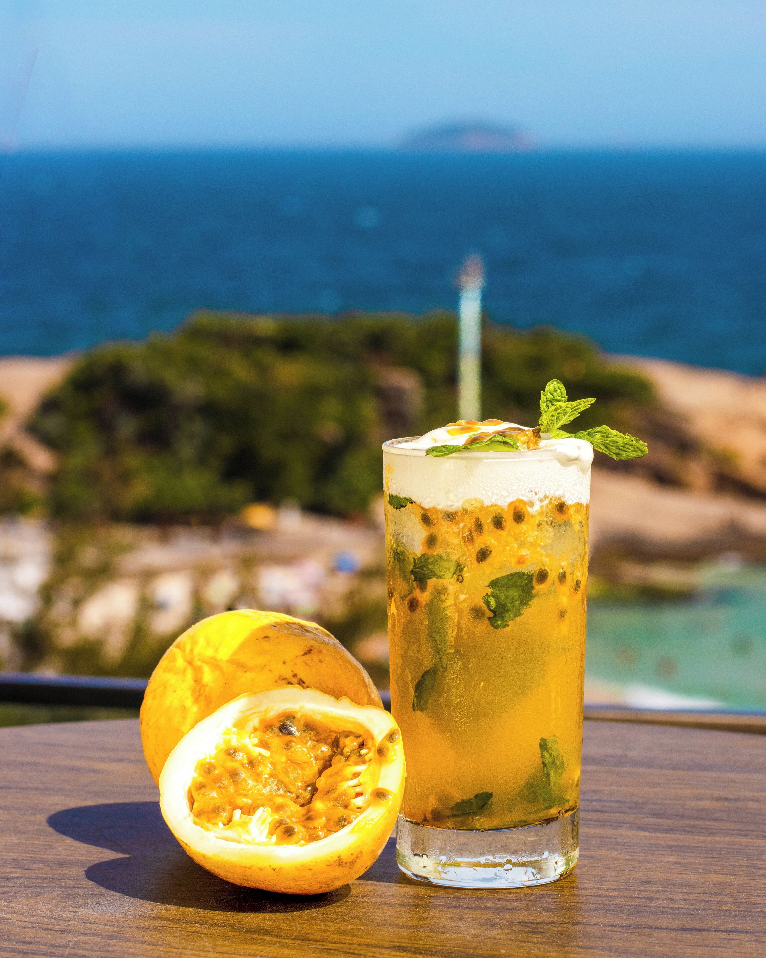 Ein Glas Eisgetränk mit Minze und einer halbierten Frucht, auf einer Holzfläche mit Blick auf die Küste.