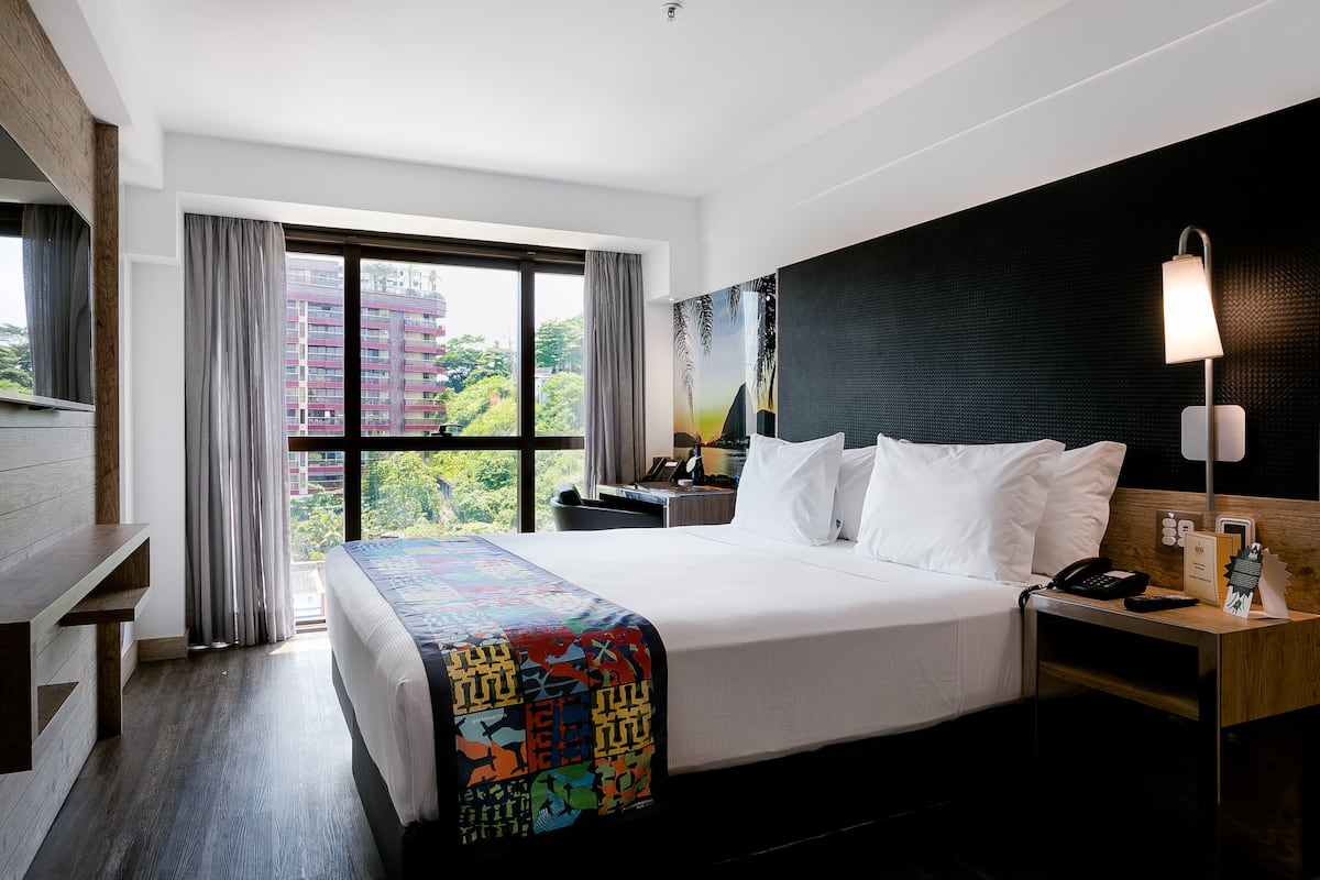 Habitación de hotel con una cama grande, un escritorio, una silla, una mesita de noche con lámpara y vistas a la ciudad desde la ventana.