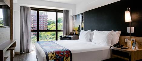 Habitación de hotel con una cama grande, un escritorio, una silla, una mesita de noche con lámpara y vistas a la ciudad desde la ventana.