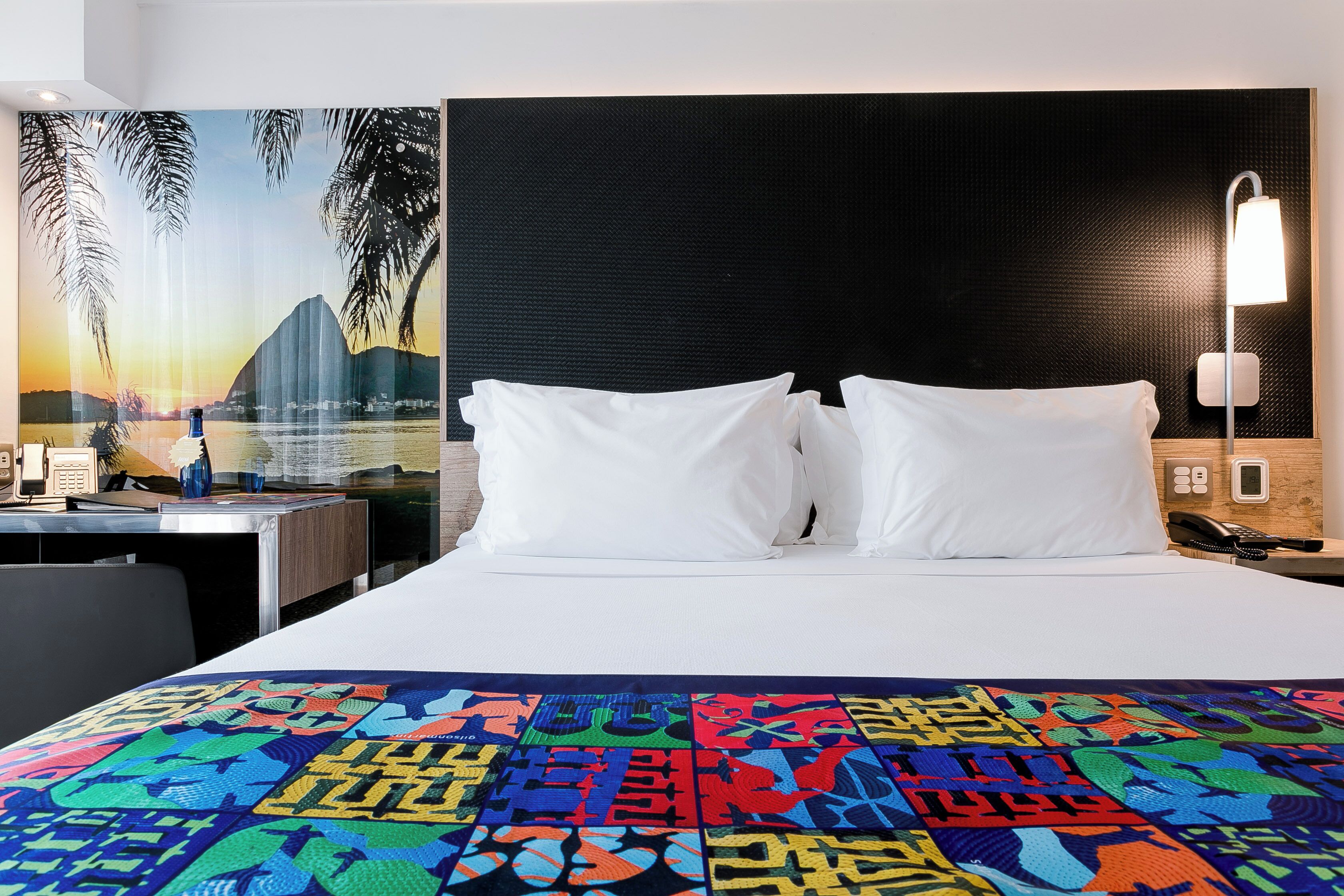 Una habitación de hotel con una cama, almohadas blancas, una manta colorida, un escritorio con un teléfono y un cuadro de un atardecer.