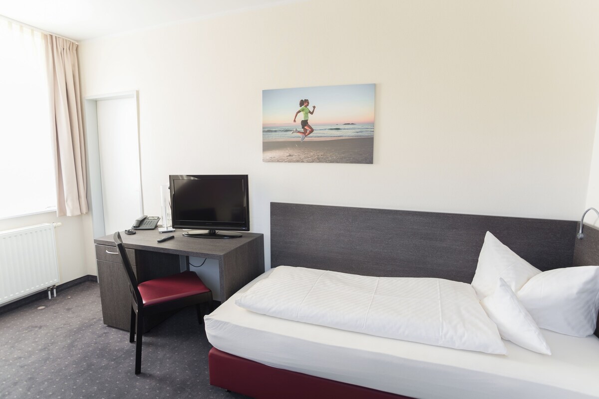 A hotel room with a bed, a desk with a computer, a chair, and a window with curtains.