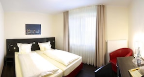 A hotel room with a double bed, a red chair, a desk with a lamp, and a painting on the wall.
