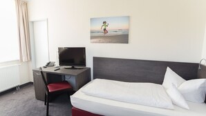 Habitación de hotel con cama, escritorio, computadora, teléfono y un cuadro de una persona corriendo en la playa.