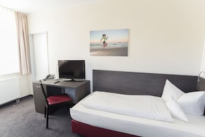 A hotel room with a bed, a desk with a computer, a chair, and a window with curtains.