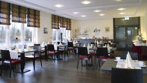 Un restaurante con varias mesas y sillas, amplias ventanas con persianas y una cocina visible al fondo.