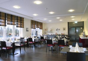 Ein Restaurant mit mehreren Tischen, Stühlen, einer Küchenzeile und großen Fenstern mit Jalousien.