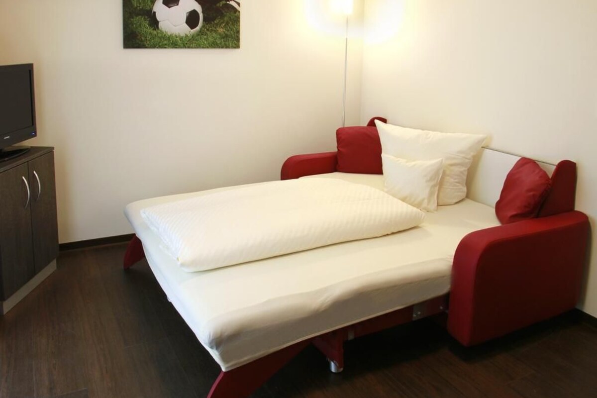 A single bed with red headboard and footboard, white bedding, and a television on a wooden cabinet.