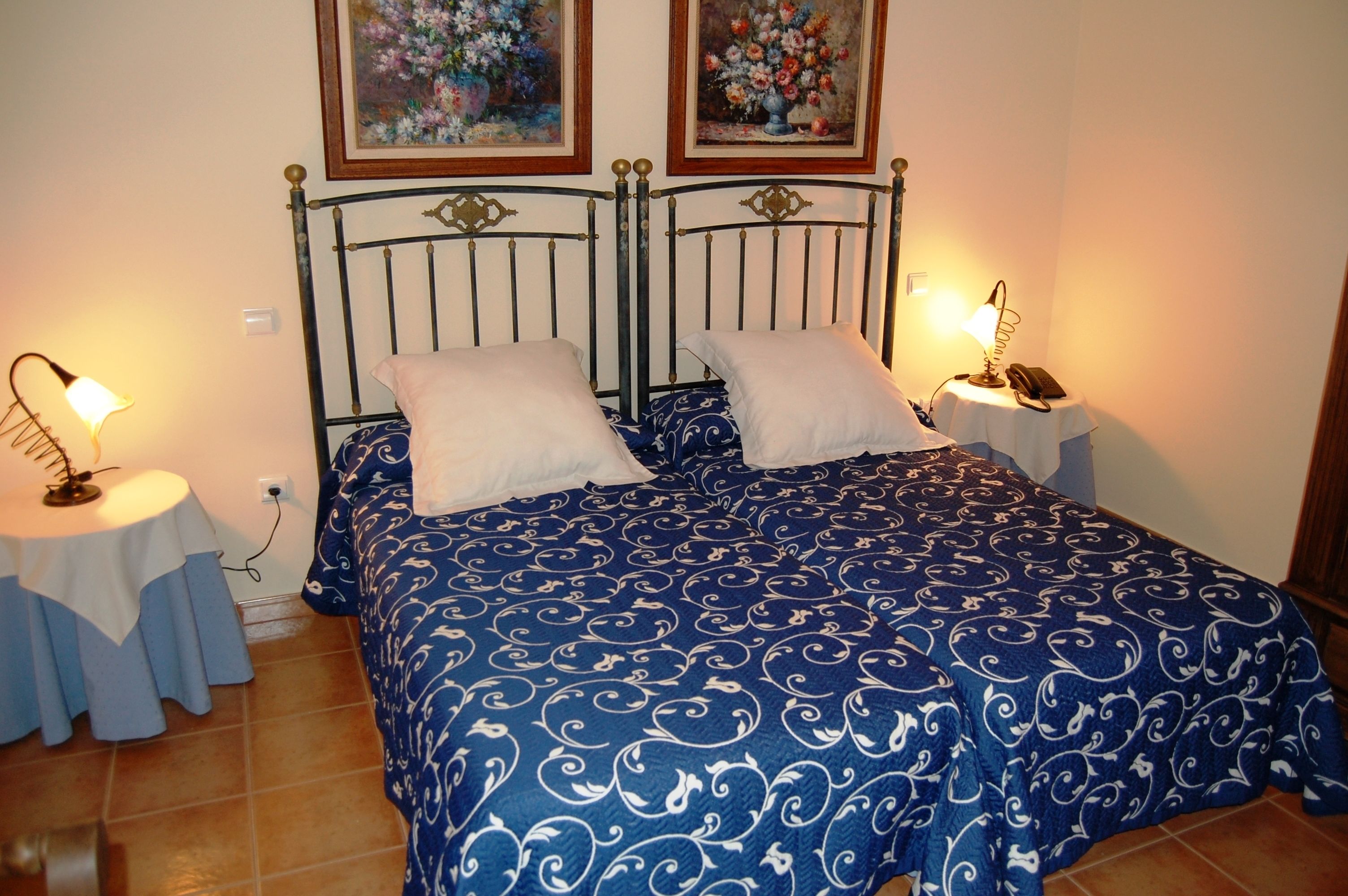 Uma cama de casal com colcha estampada em azul e branco, duas almofadas brancas, cabeceira de metal e duas mesas de cabeceira com luminárias.