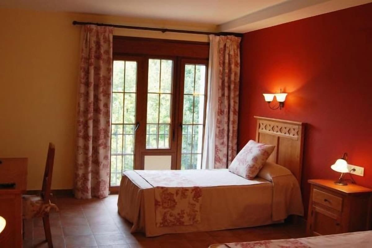 Un dormitorio con cama, escritorio, silla, mesita de noche, lámpara y ventana con cortinas.