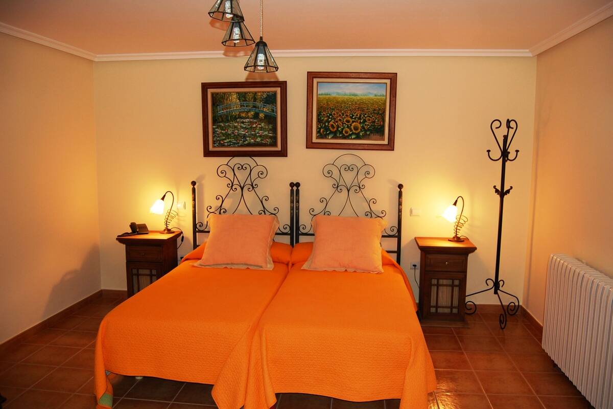 A double bed with orange bedding, two bedside tables with lamps, and two framed pictures on the wall.