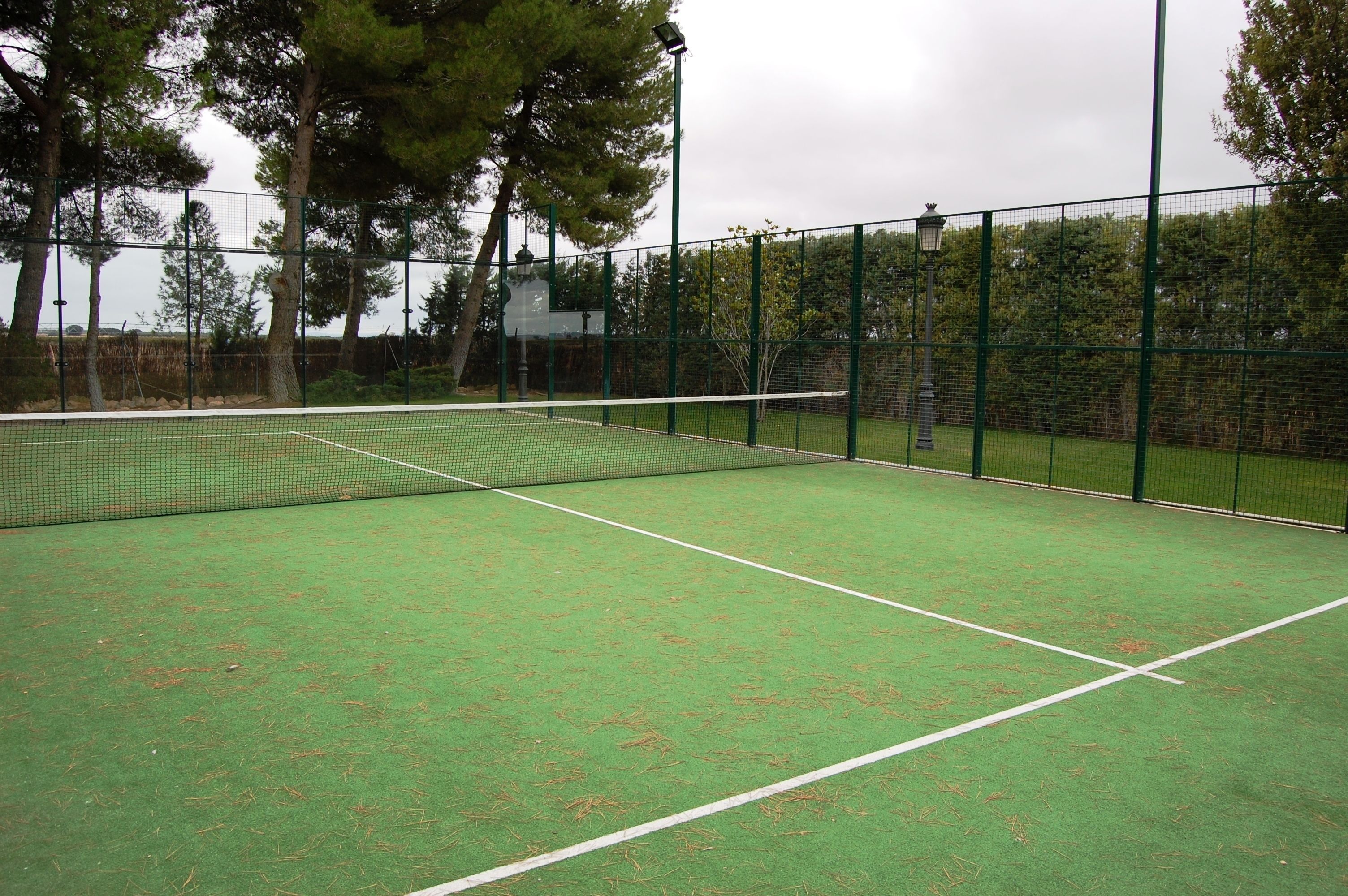 A tennis court with a net, surrounded by a green fence and trees.
