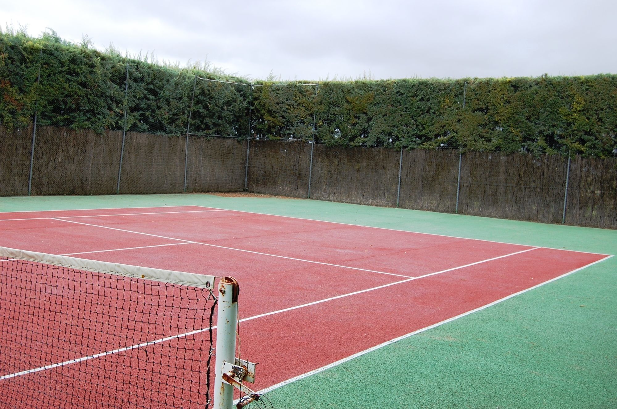 Ein Hallen-Tennisplatz mit roter Spielfläche und einem umlaufenden Grünbereich, begrenzt von einem Maschendrahtzaun und einer Hecke.
