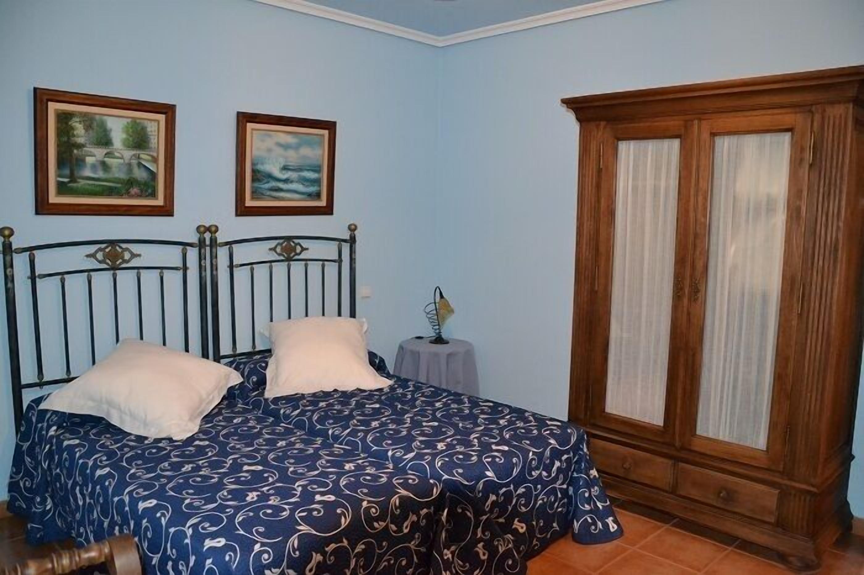 A bedroom with a double bed, two framed pictures on the wall, a small round table, and a wooden wardrobe.