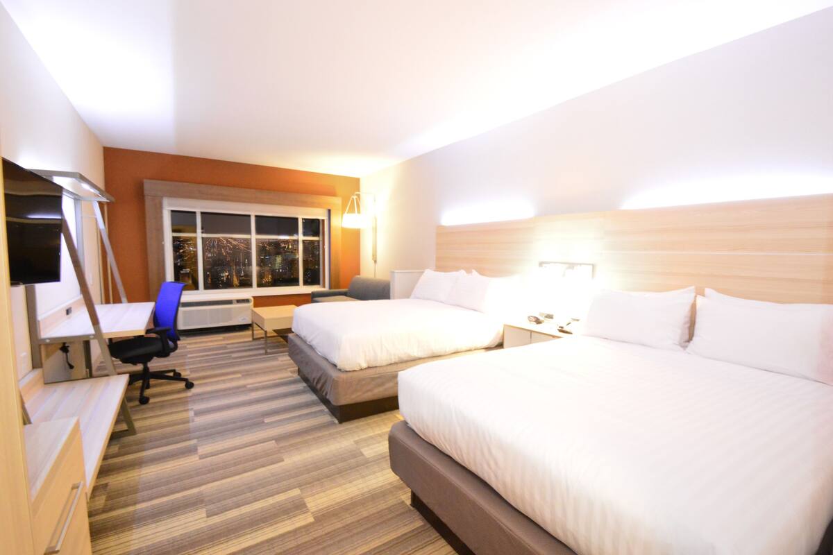 Ein Hotelzimmer mit zwei Betten, einem Schreibtisch, einem Sessel, einem Fernseher und einem Fenster mit Blick auf die Stadt.