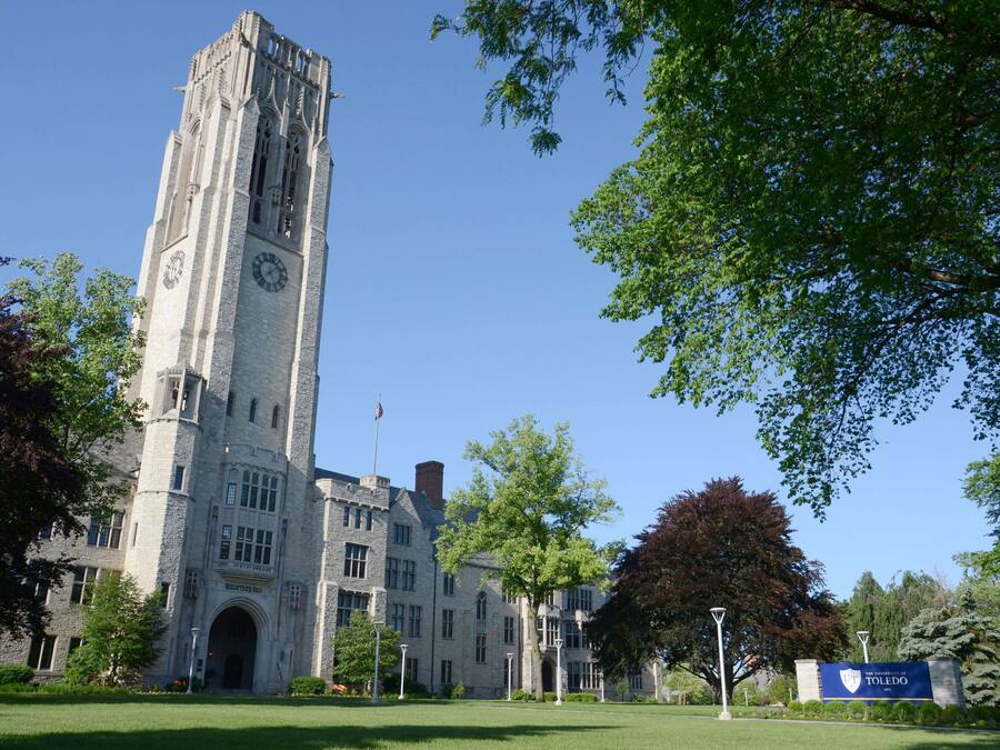 A historic stone building with a clock tower, surrounded by trees and a well-maintained lawn.
