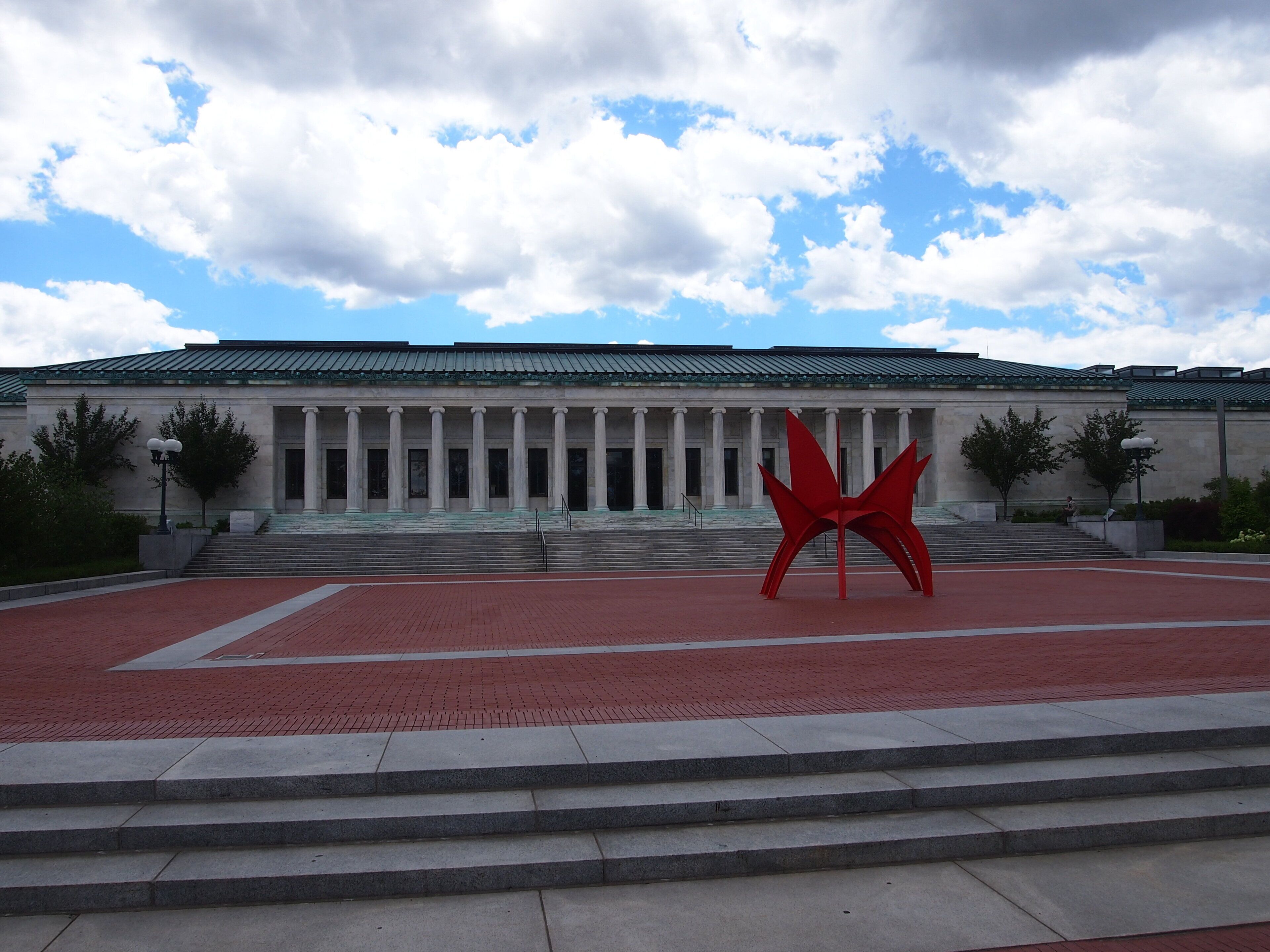 A building with a classical architectural style, featuring a portico with columns, a red brick pathway leading to the entrance, and a large red sculpture in the foreground.