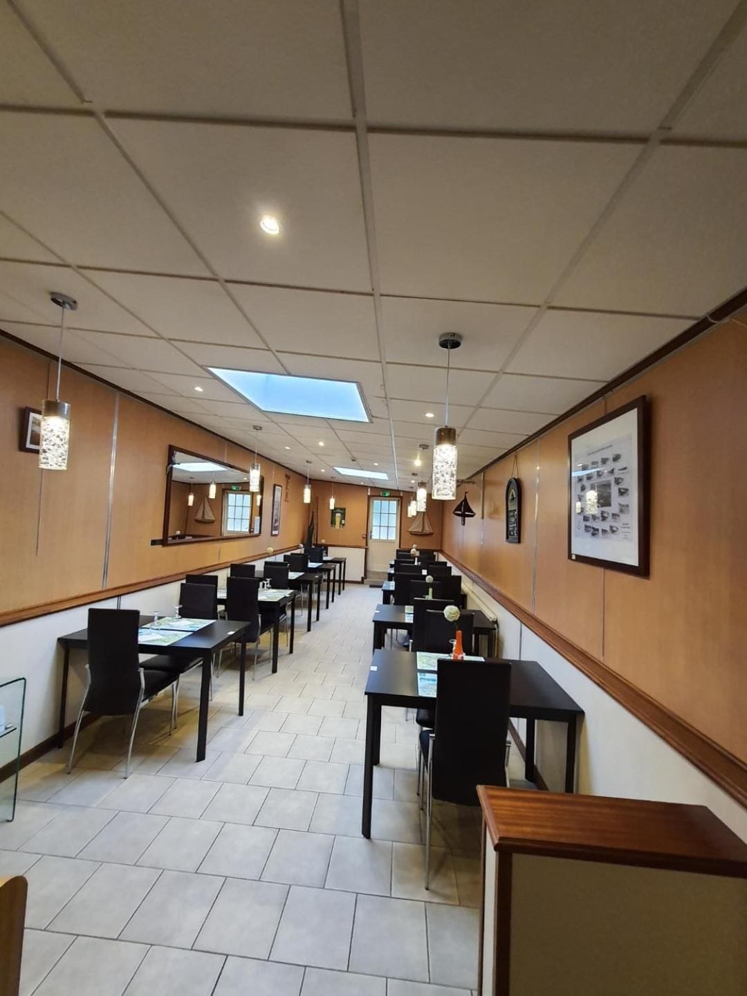 Une salle à manger d’hôtel avec de nombreuses tables et chaises, une verrière et des tableaux encadrés aux murs.