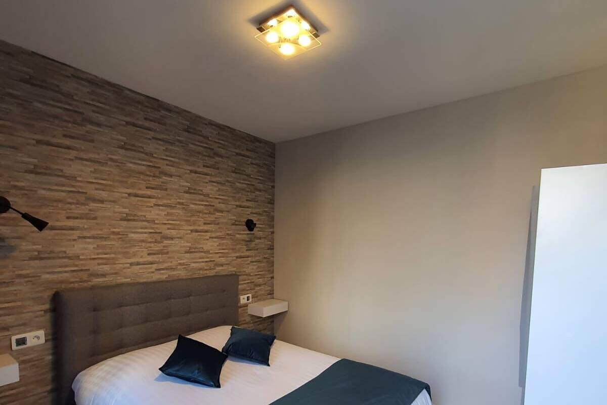 A modern bedroom with a wooden wall, a bed with a dark headboard, two pillows, a bedside table with a lamp, and a ceiling light fixture.