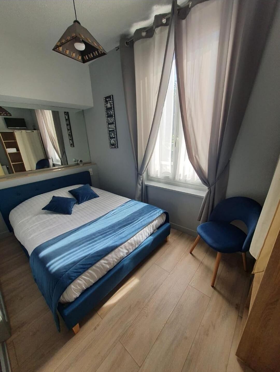 Een slaapkamer met een bed, een stoel, een raam met gordijnen, een spiegel en een aan de muur hangend schilderij.