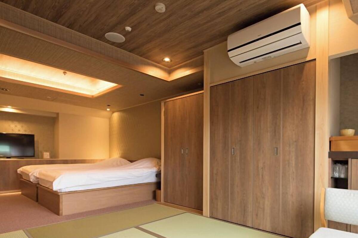 A hotel room with a bed, a wooden wardrobe, a small table, a TV, and an air conditioning unit.