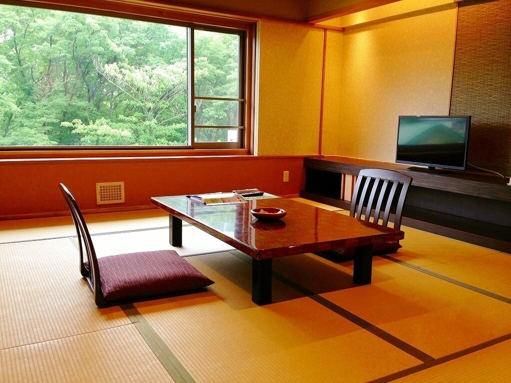 A traditional Japanese room with tatami flooring, a low table, a cushioned bench, a television, and a window with a view of greenery.