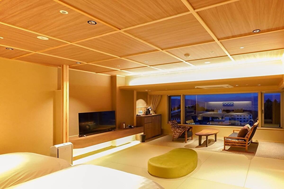A hotel room with a bed, a TV, a desk, a chair, a sofa, and a large window with a view.