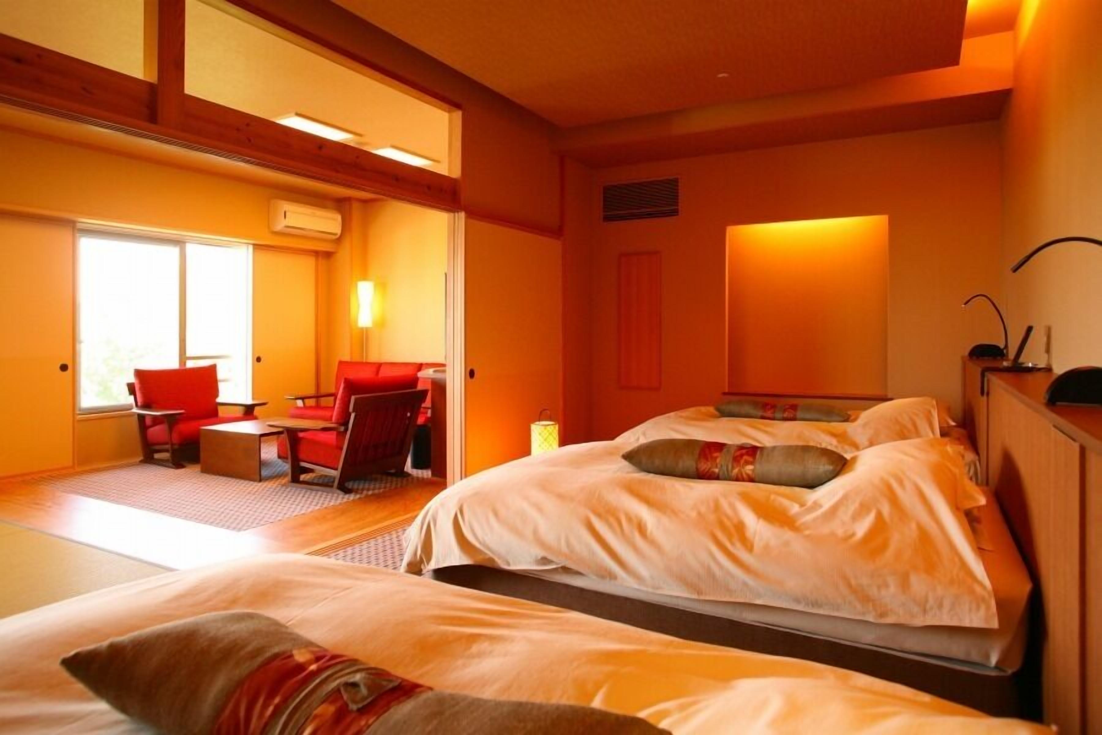 A hotel room with two beds, a sitting area with a red chair, and a large window.