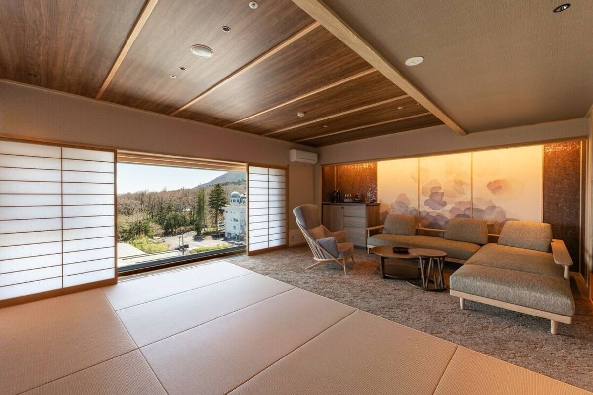 A modern living room with tatami flooring, a sofa, a chair, and a view of a mountain.