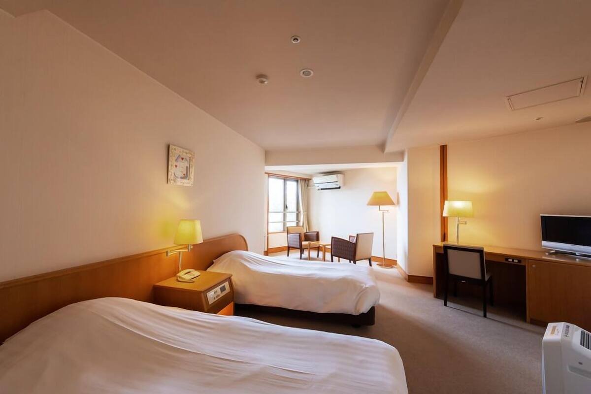 A hotel room with two beds, a desk, a chair, a TV, and a lamp.