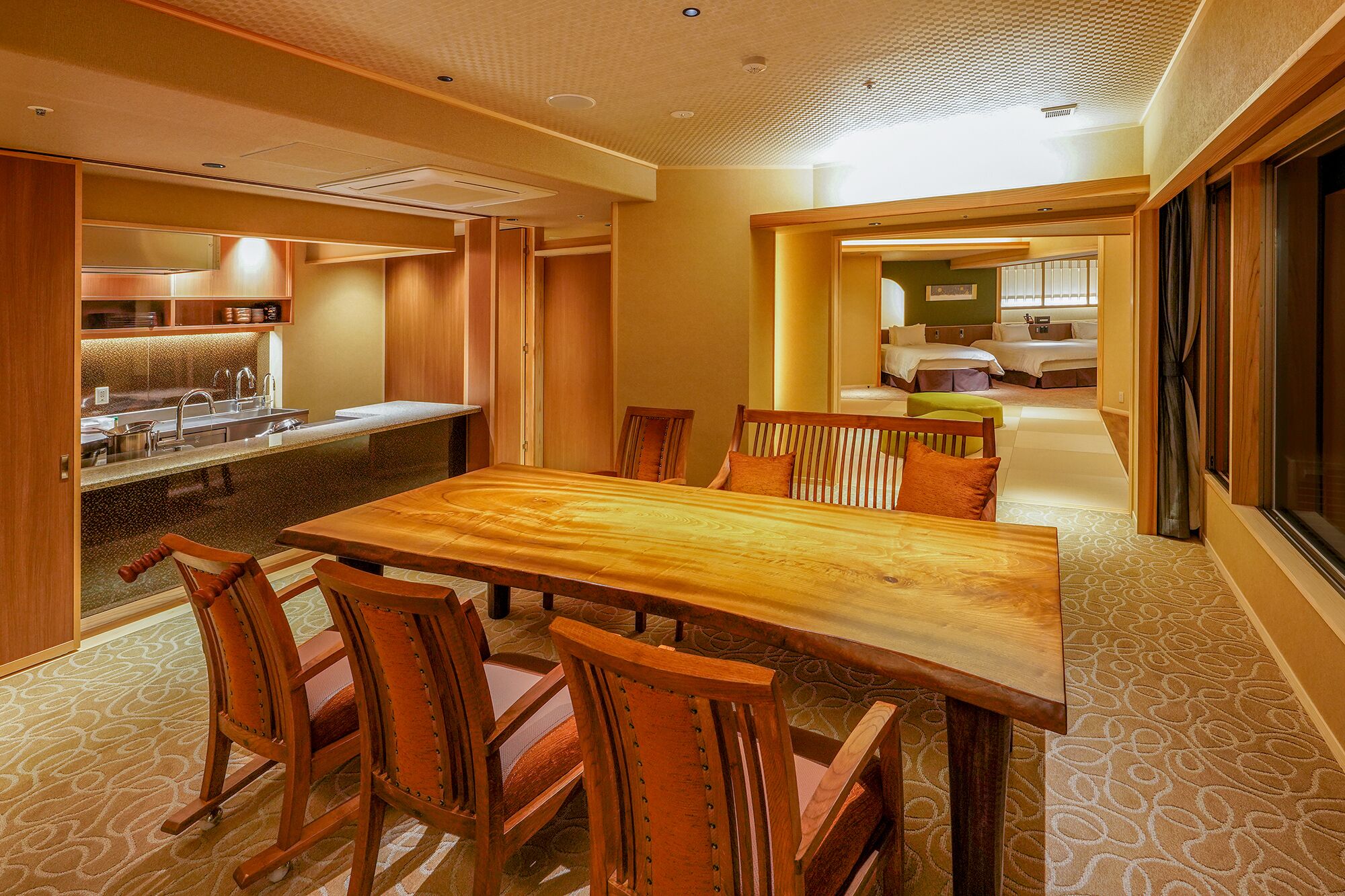 A hotel room with a wooden dining table and chairs, a kitchen area with a sink and cabinets, and a view of the adjacent bedroom with two beds.