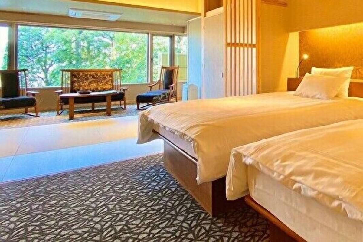 A hotel room with a large bed, a seating area with chairs, a view of greenery outside, and a patterned floor.
