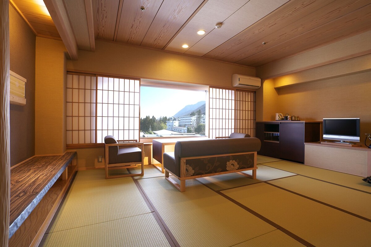 A traditional Japanese room with tatami flooring, a low table, a sofa, a TV, and a view of mountains.