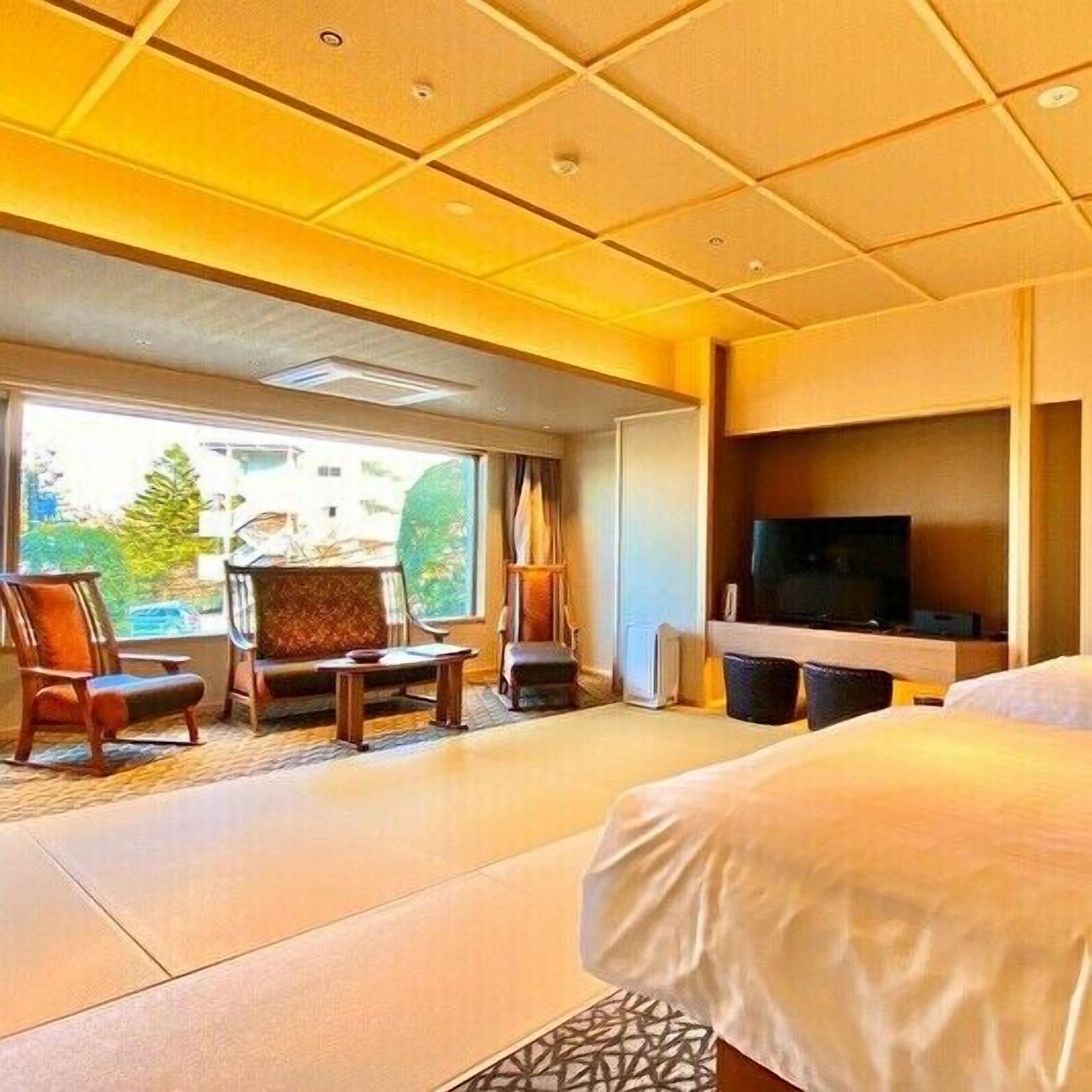 A hotel room with a bed, a TV, a seating area with chairs, and a large window.