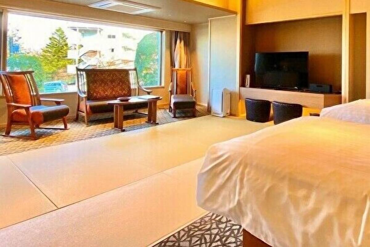 A hotel room with a bed, a TV, a seating area with chairs, and a large window.
