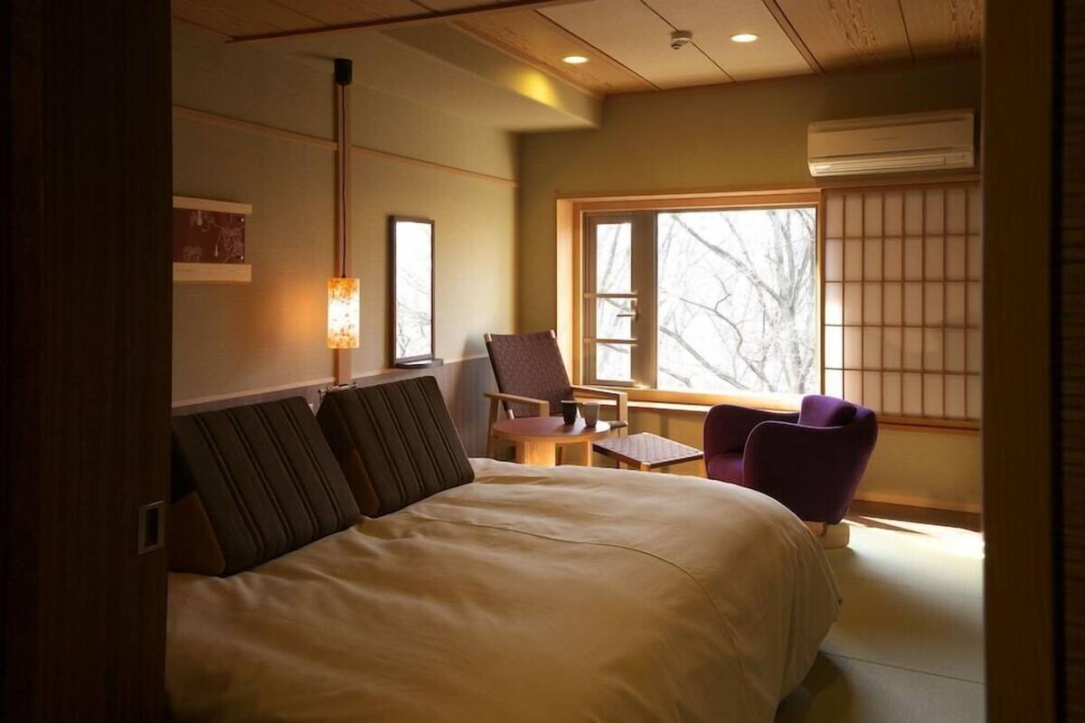 A hotel room with a bed, a chair, a small table, a window with a view, and an air conditioning unit.