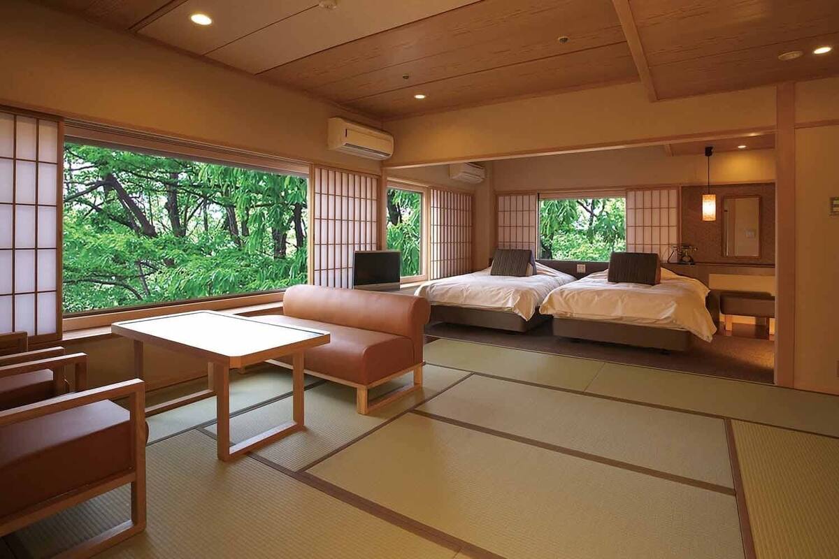 A traditional Japanese-style room with tatami flooring, a low table, a sofa, two beds, and large windows with a view of greenery.