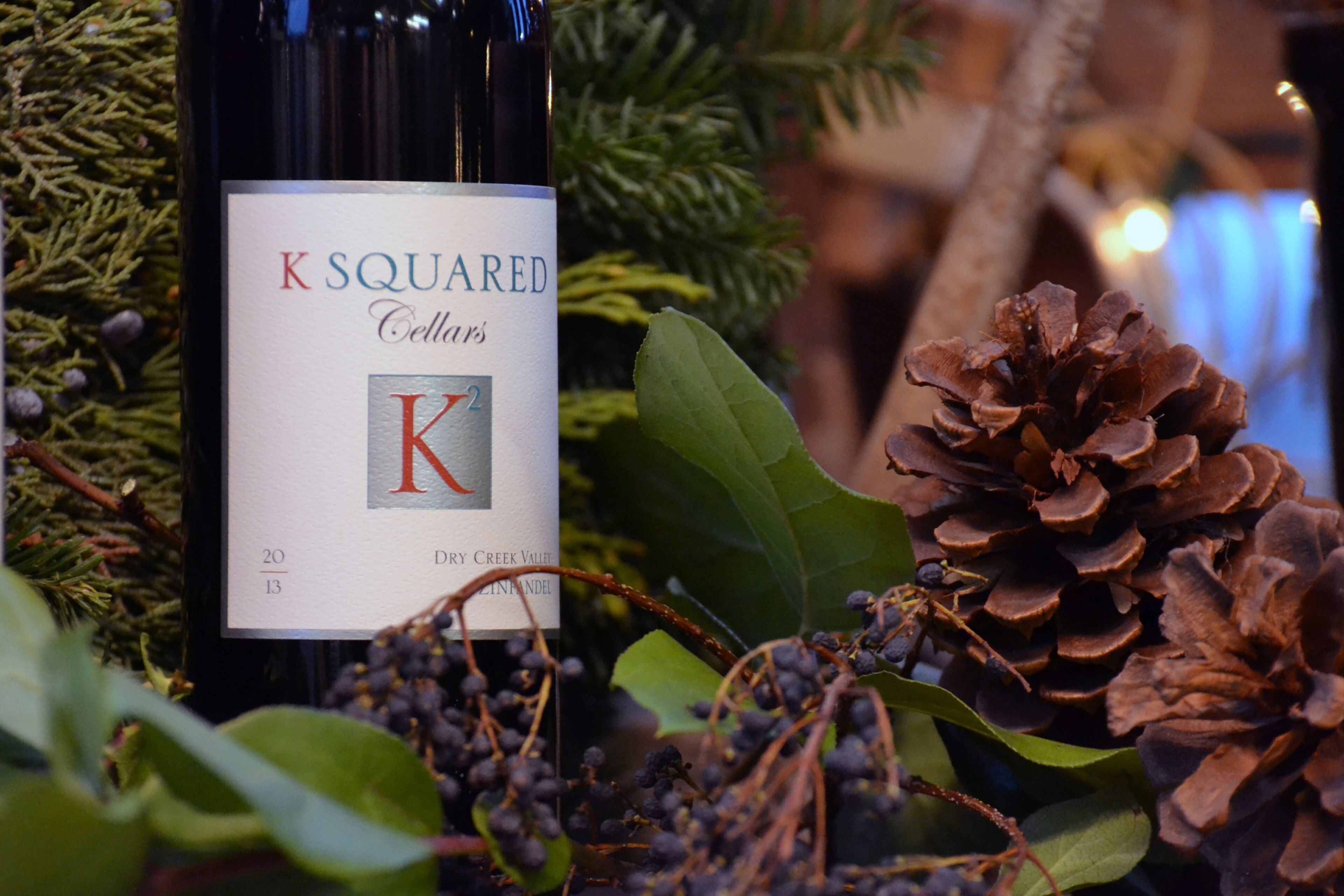 A bottle of K Squared Cellars wine with a label, surrounded by pine cones and greenery.