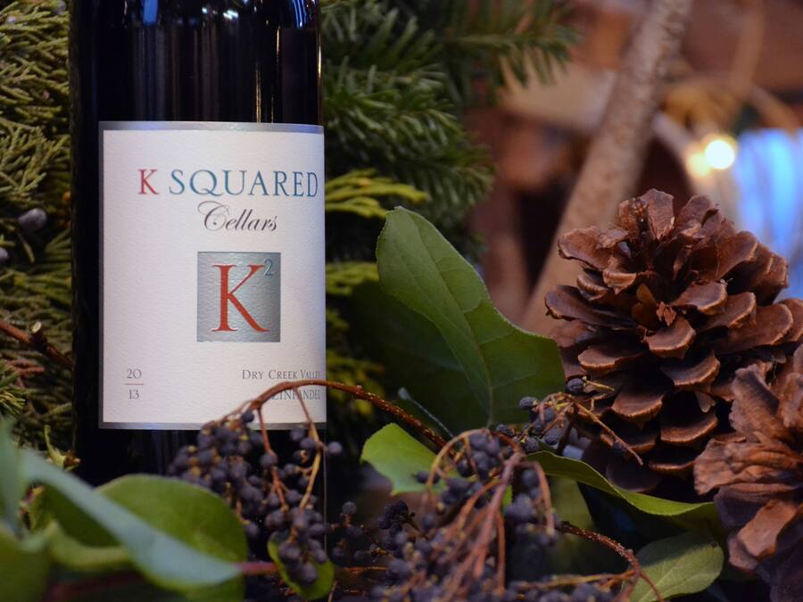 A bottle of K Squared Cellars wine with a label, surrounded by pine cones and greenery.