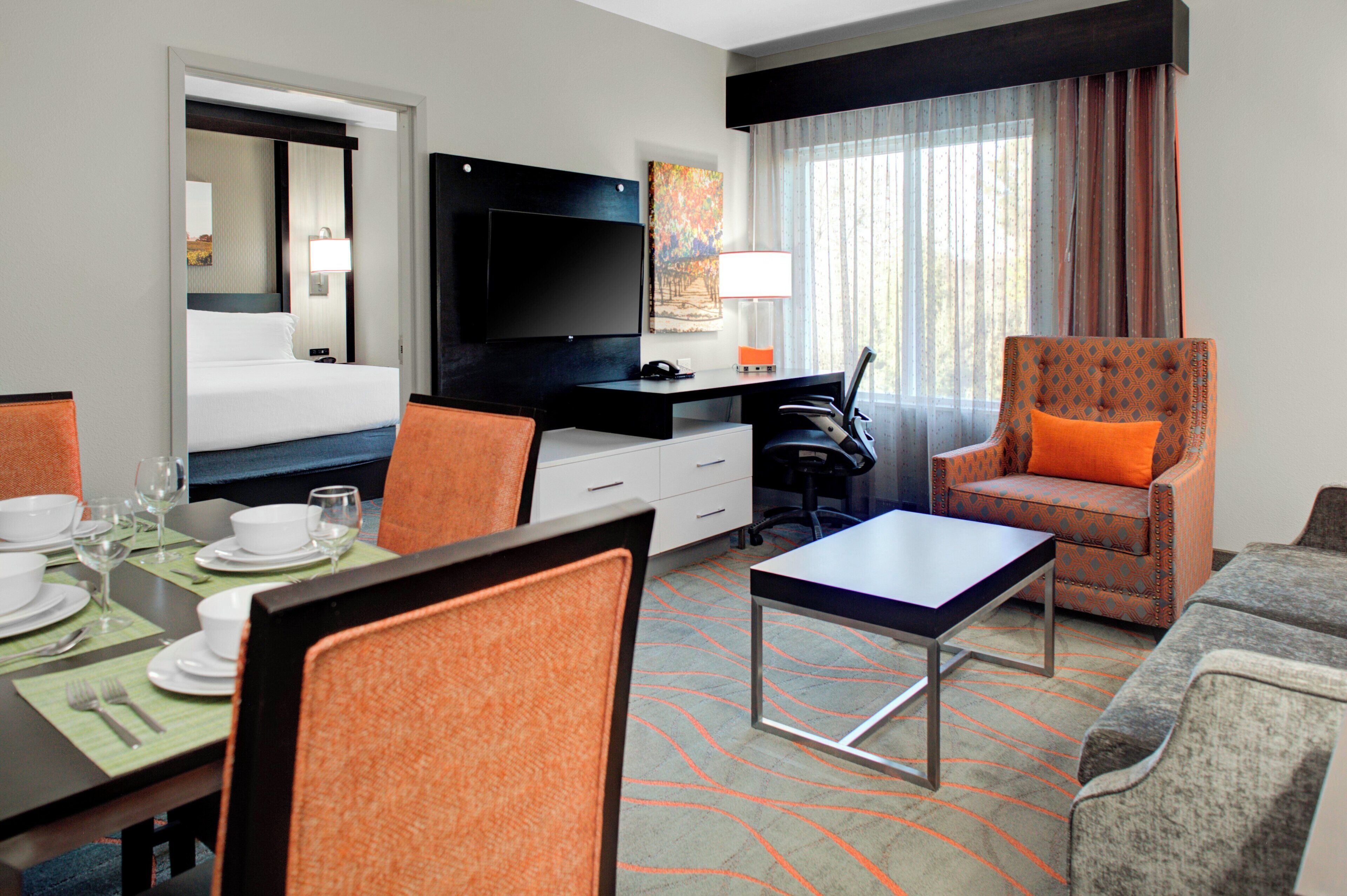 A hotel room with a bed, a desk with a TV, a dining area with a table and chairs, a sofa, and a window with curtains.