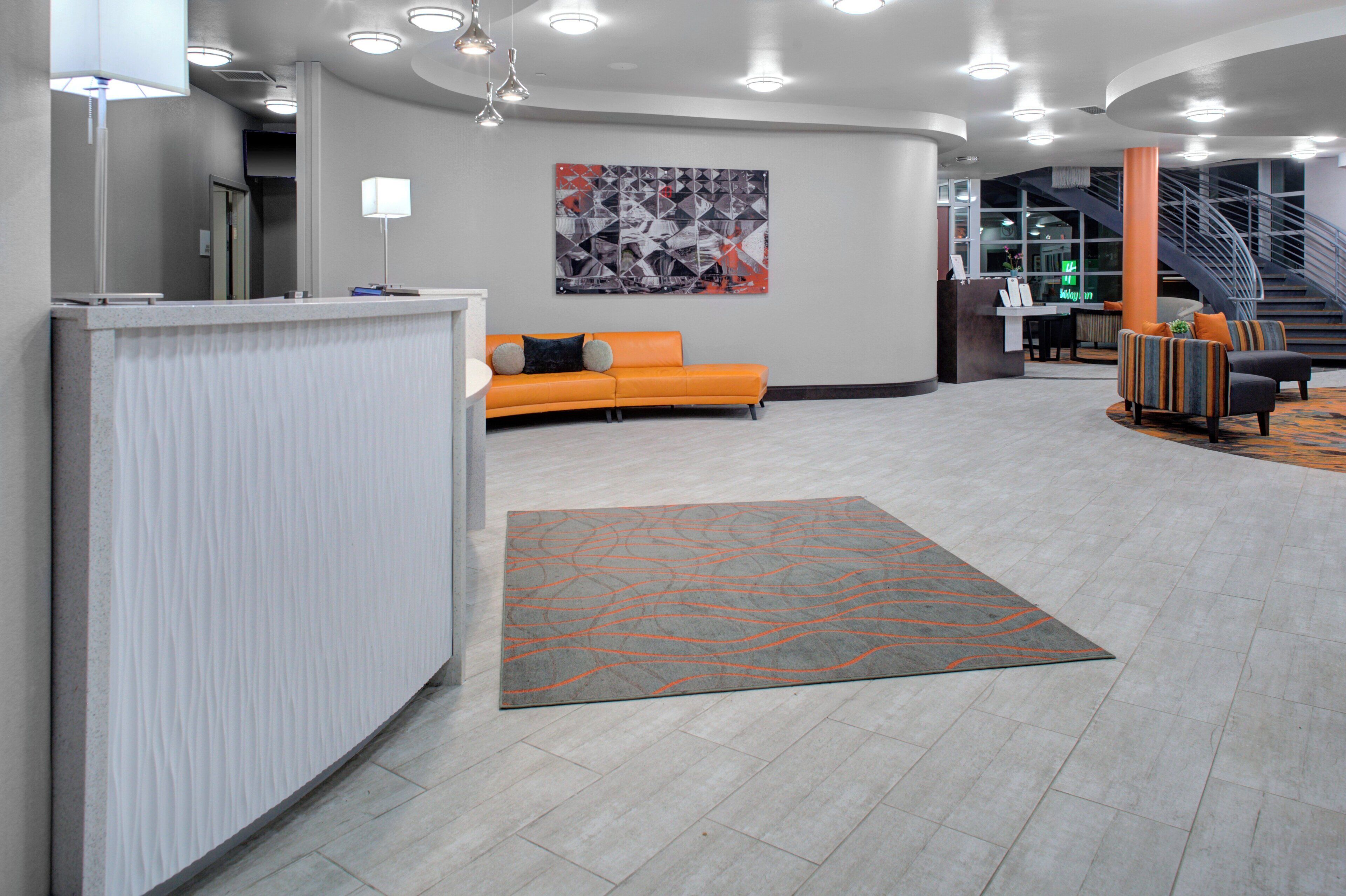 En moderne lobby med en reception, et siddeområde med en orange sofa, et mønstret tæppe og en trappe i baggrunden.