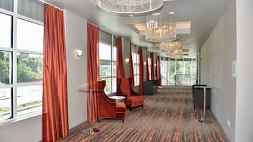 A hotel corridor with a patterned carpet, red upholstered seating, and large windows with curtains.