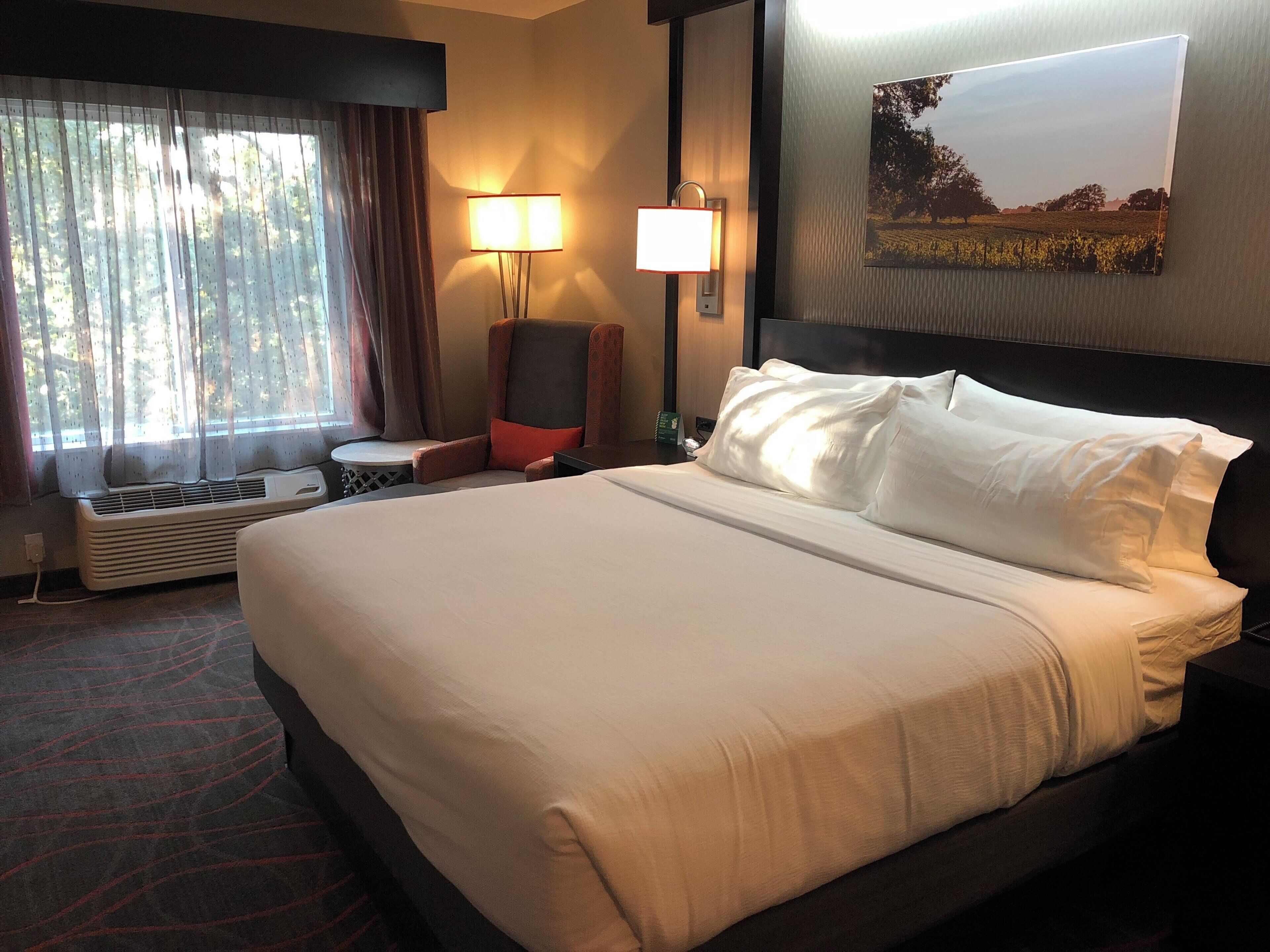 A hotel room with a large bed, a chair, a table, and a wall-mounted lamp.
