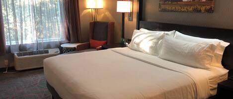 A hotel room with a large bed, a chair, a table, and a wall-mounted lamp.