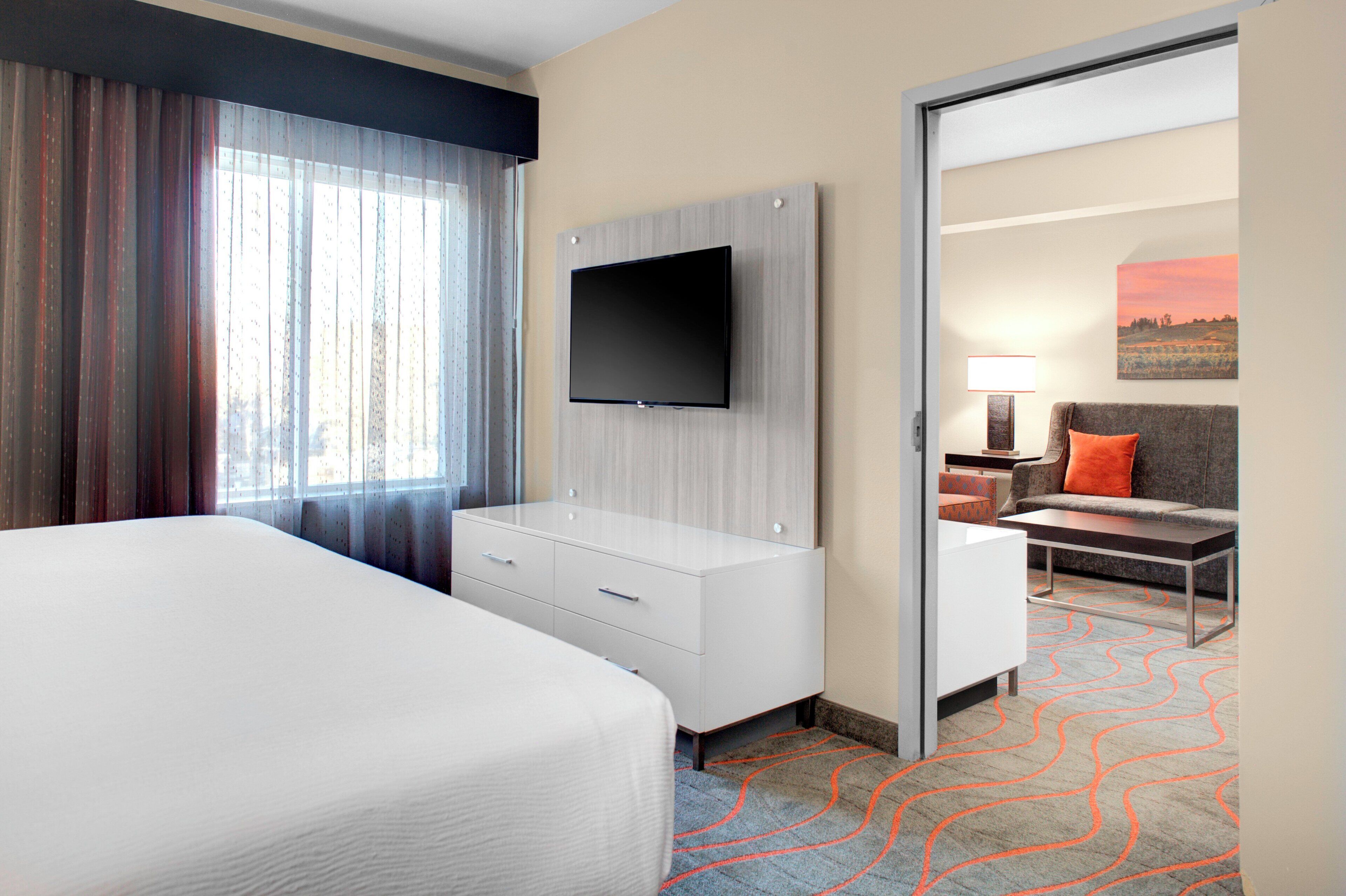 A hotel room with a bed, a TV mounted on the wall, a desk with a chair, and a view through an open door.