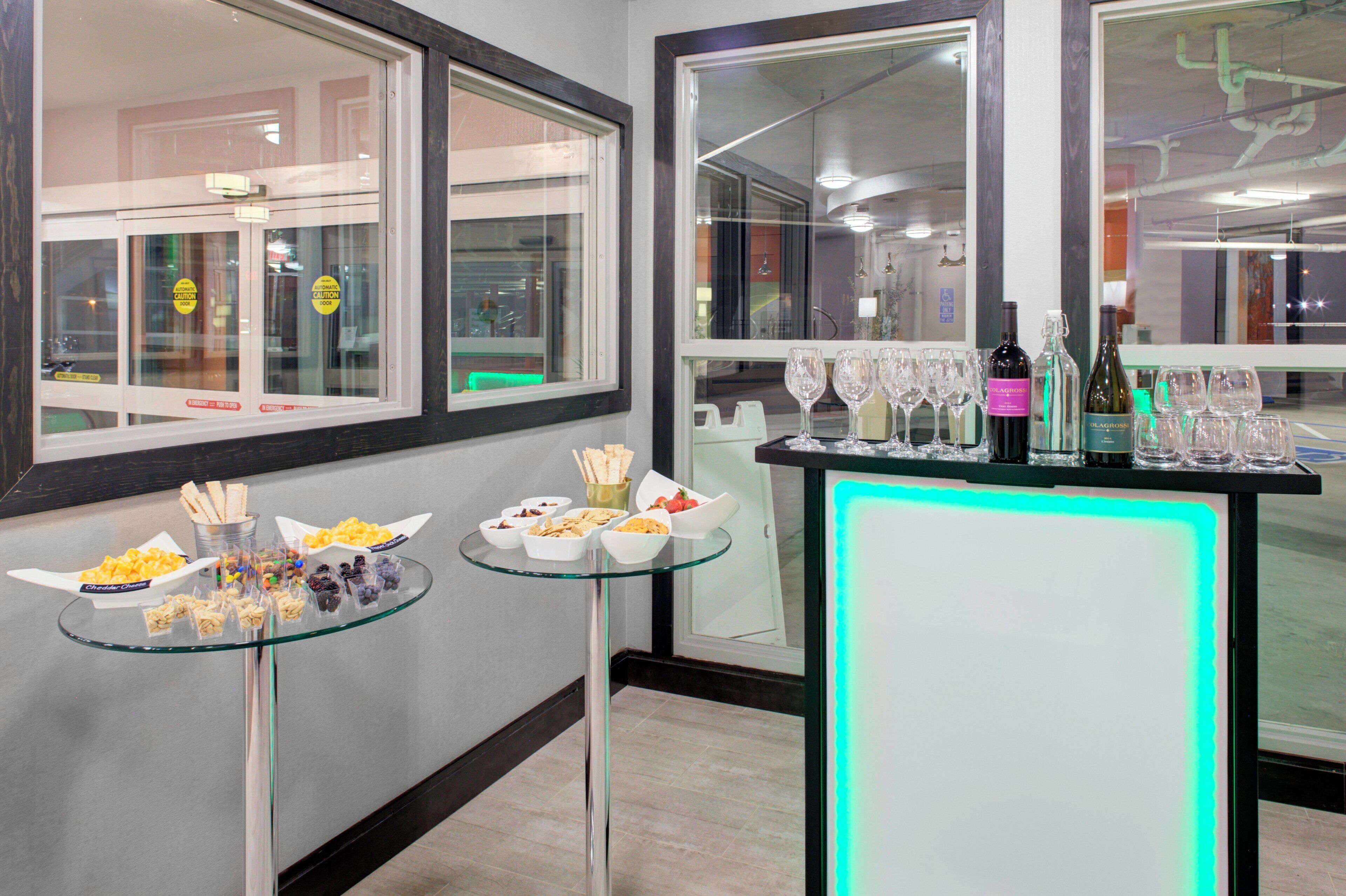 Quarto com mesa de vidro exibindo petiscos e área de bar com copos e garrafas de vinho.