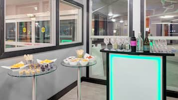 Quarto com mesa de vidro exibindo petiscos e área de bar com copos e garrafas de vinho.