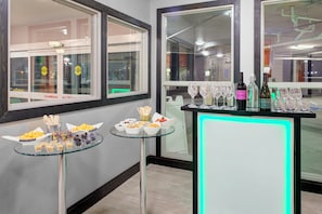 A room with a glass table displaying snacks and a bar area with wine glasses and bottles.
