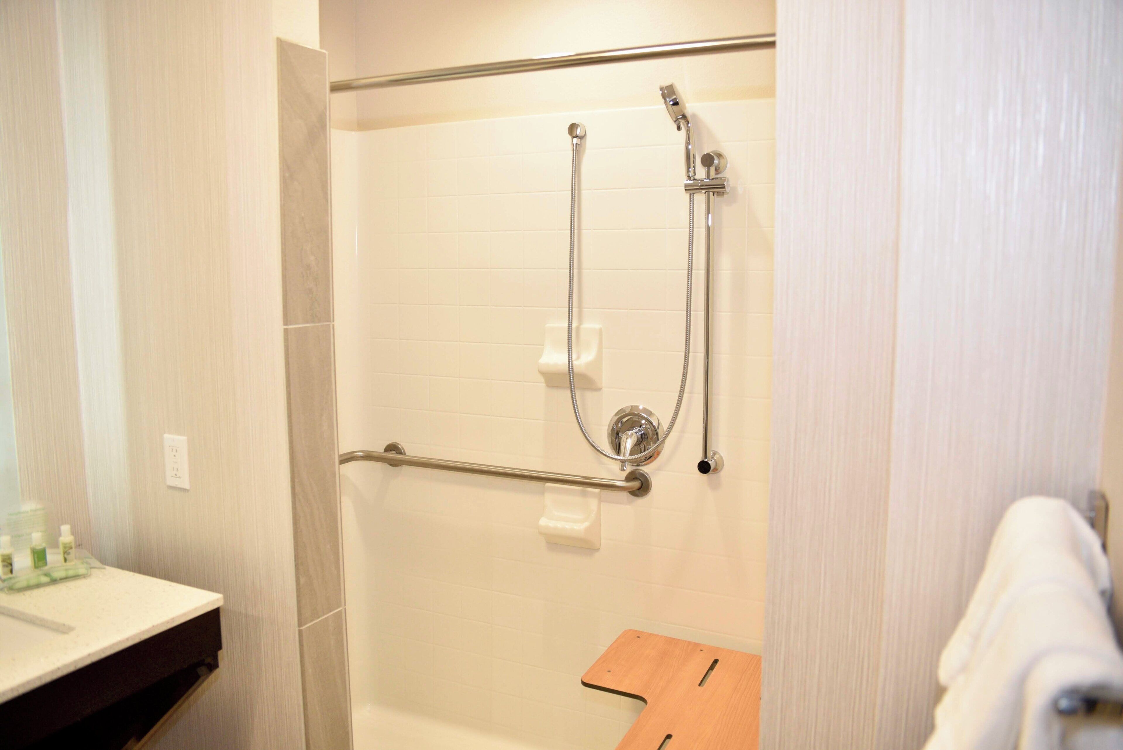 A shower with a grab bar, a white tiled wall, and a wooden bench.